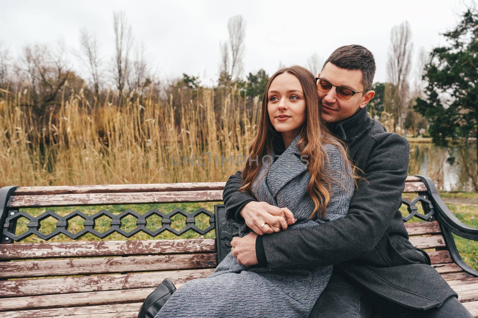 A man and woman are sitting closely on a park bench, surrounded by autumn foliage and a serene lake. They share a tender moment, enjoying each other's company.