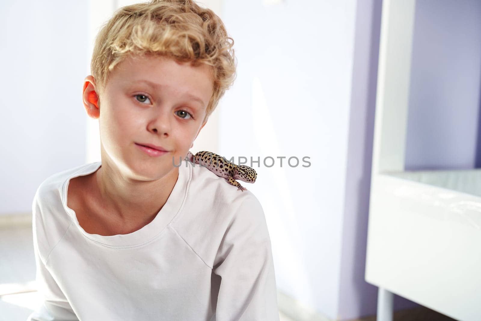 A cheerful young boy sits comfortably on the floor, smiling as a leopard gecko rests on his shoulder. The room is well-lit and has light-colored walls.