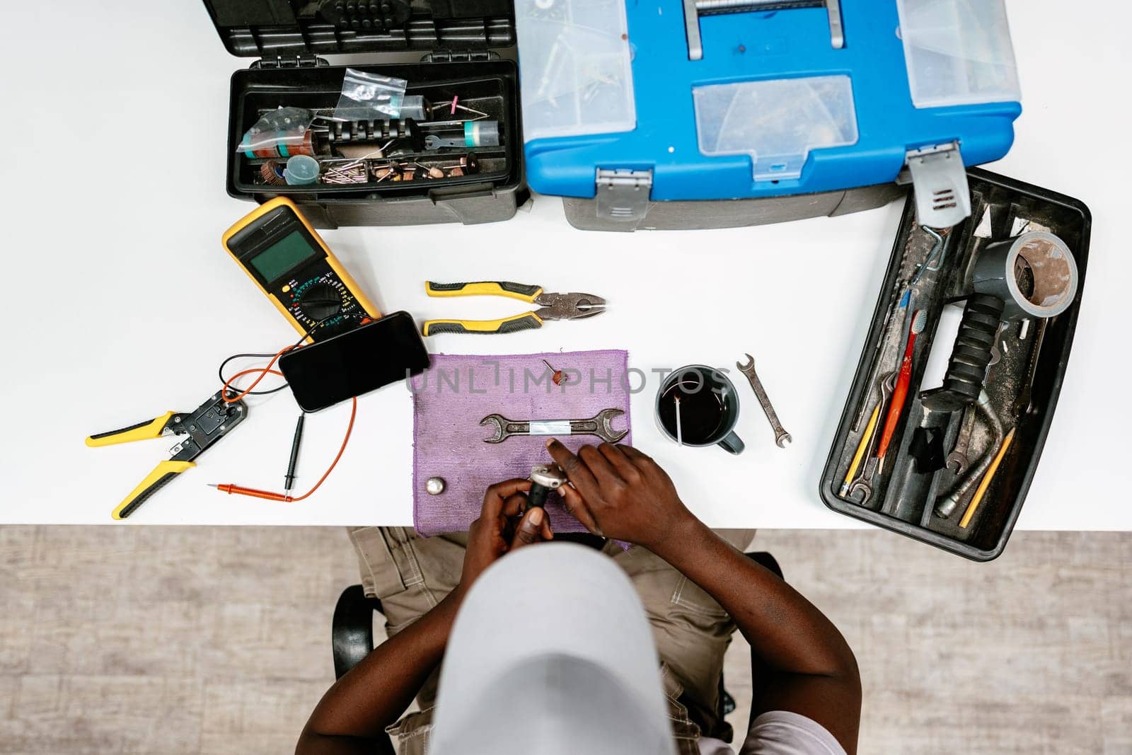 A technician works on an electronic device, using various tools like pliers and wrenches on a workbench. The area is organized with tools and a smartphone. Daylight fills the workspace.