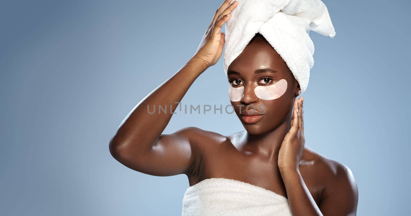A young woman with a towel wrapped around her hair applies an under-eye mask in a simple studio. The soft blue background enhances her natural beauty.