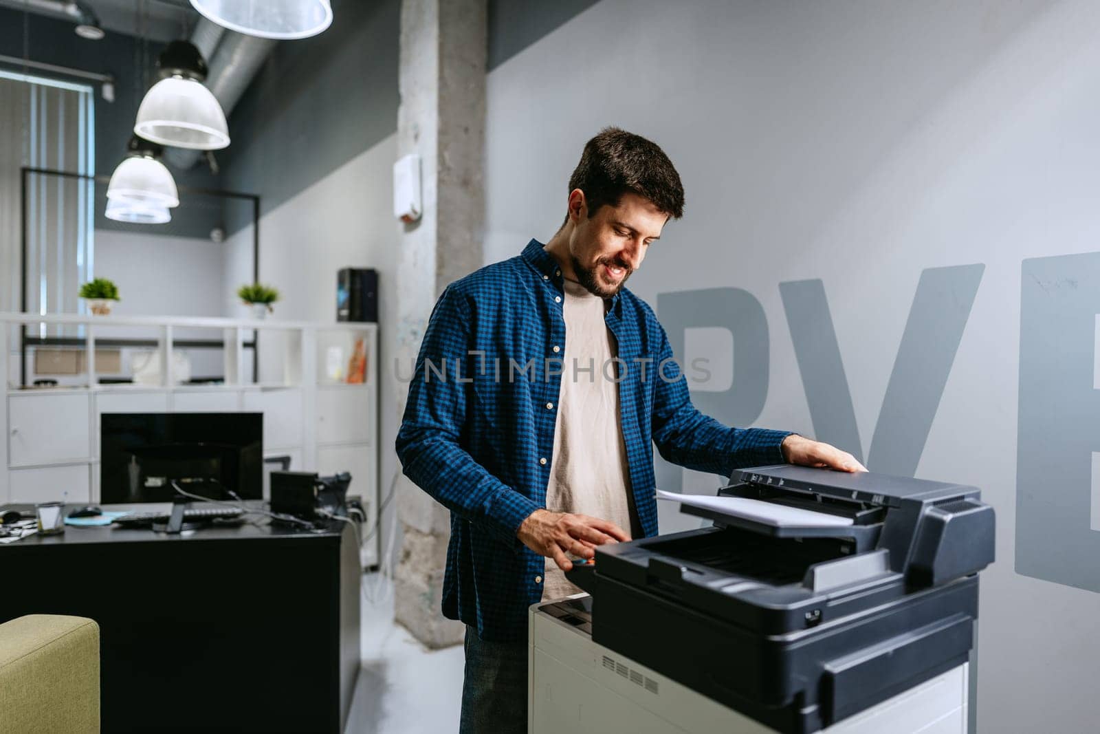 A man in a blue checkered shirt uses a printer in a contemporary office space filled with natural light. The atmosphere is relaxed, emphasizing productivity and collaboration.