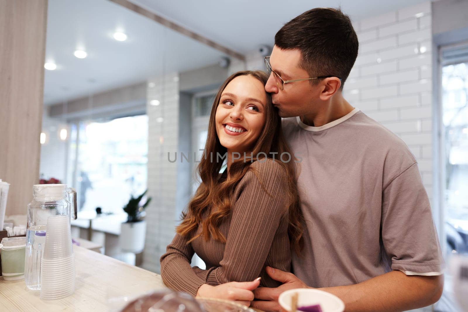 A couple enjoys an intimate moment in a charming cafe. The man leans in to kiss the woman on the cheek, who beams with happiness. Soft natural light pours in through the windows.