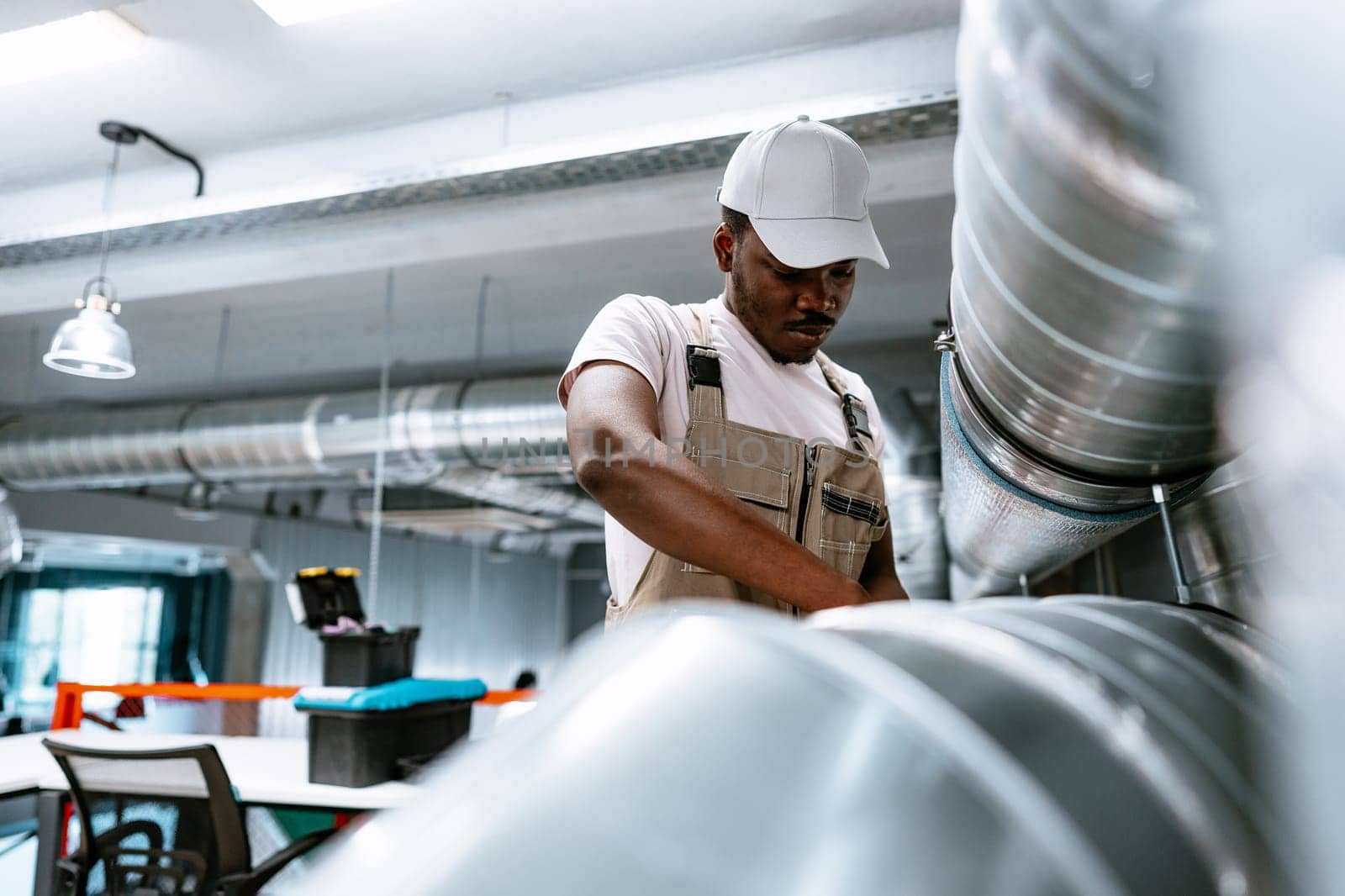 A worker in beige overalls and a white cap is focused on repairing equipment in a well-lit workshop. The environment features metal ducting and workstations.