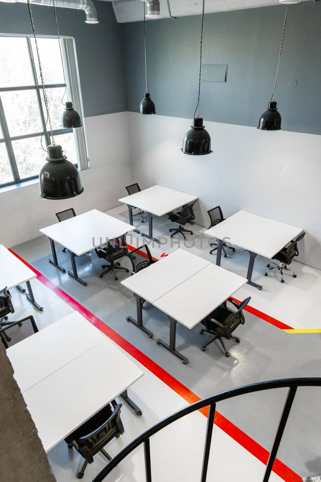A spacious office interior showcases multiple white tables and ergonomic chairs arranged in a functional layout. The bright space is illuminated by large windows and hanging lights.
