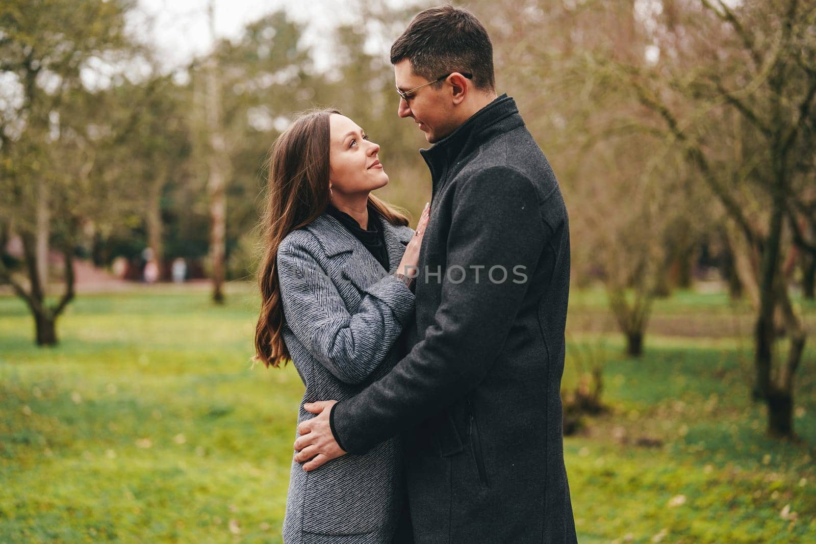 A couple stands close together in a green park, gazing into each other's eyes. They share a warm connection, surrounded by nature's beauty on an overcast day.