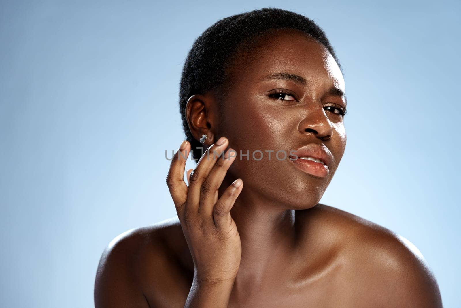 A woman with a short hairstyle stands against a light blue backdrop. She exudes confidence as she subtly touches her face, showcasing her features with a poised expression.