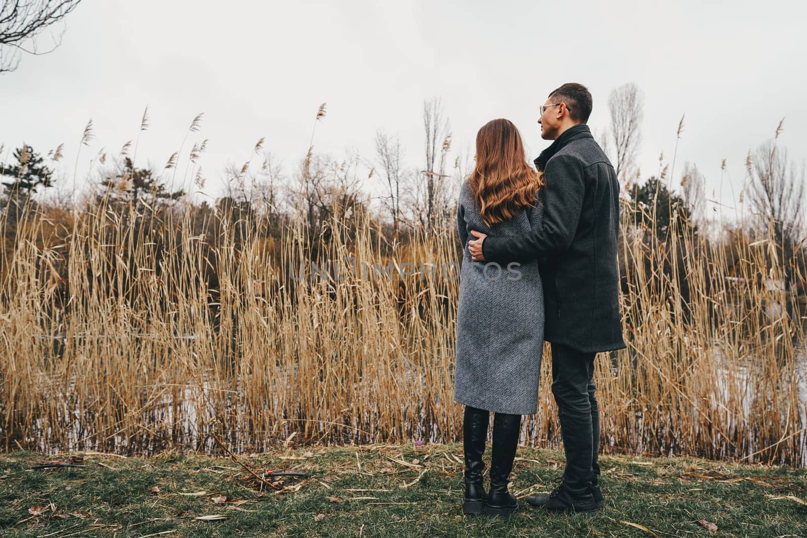 Two individuals stand close together, embracing gently as they gaze towards a serene landscape. Tall grasses sway in the wind near a calm body of water, creating a peaceful atmosphere.
