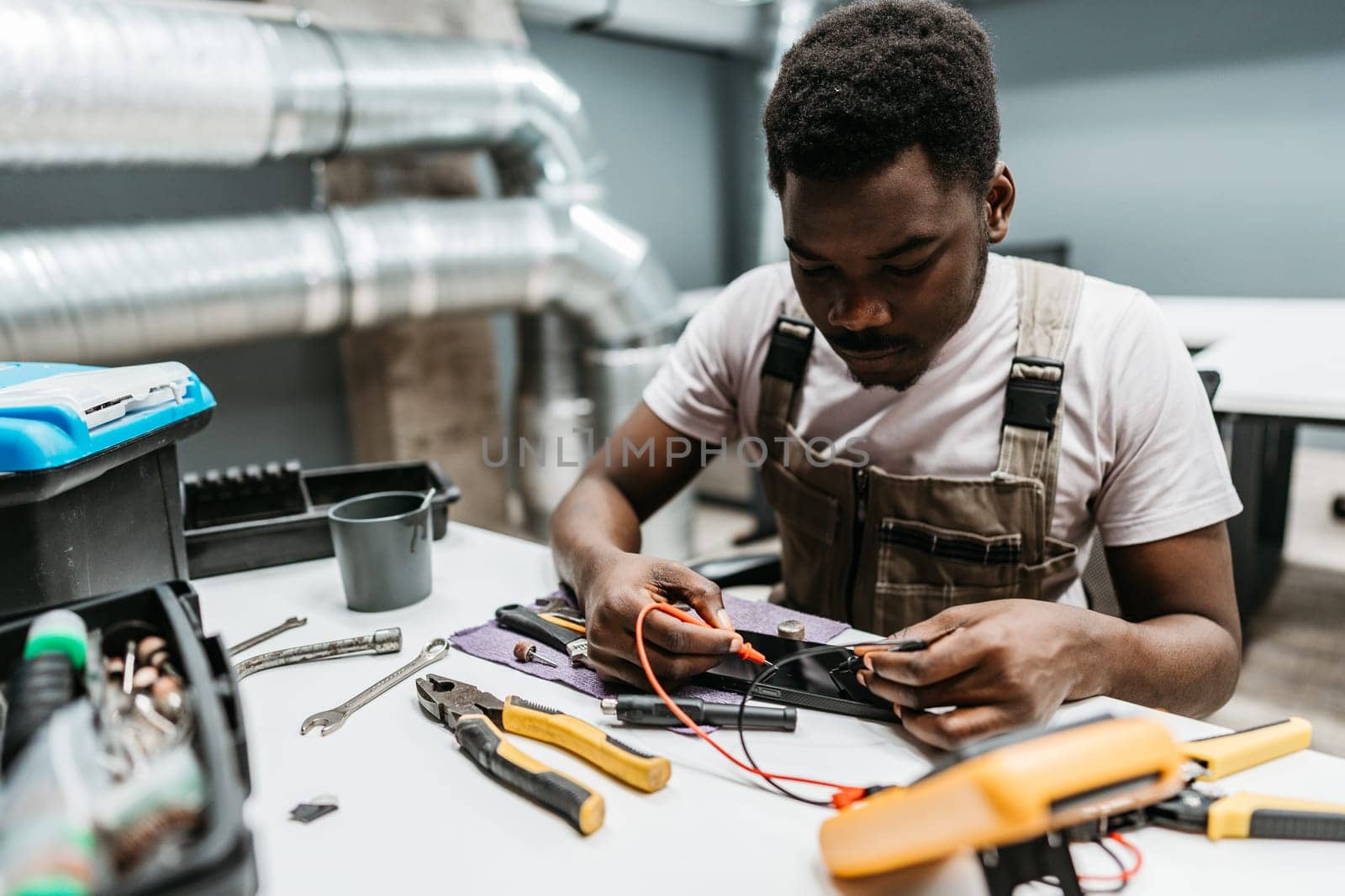 A young technician works diligently on electrical wiring in a workshop. Surrounded by tools and equipment, he carefully connects wires while seated at a workbench.