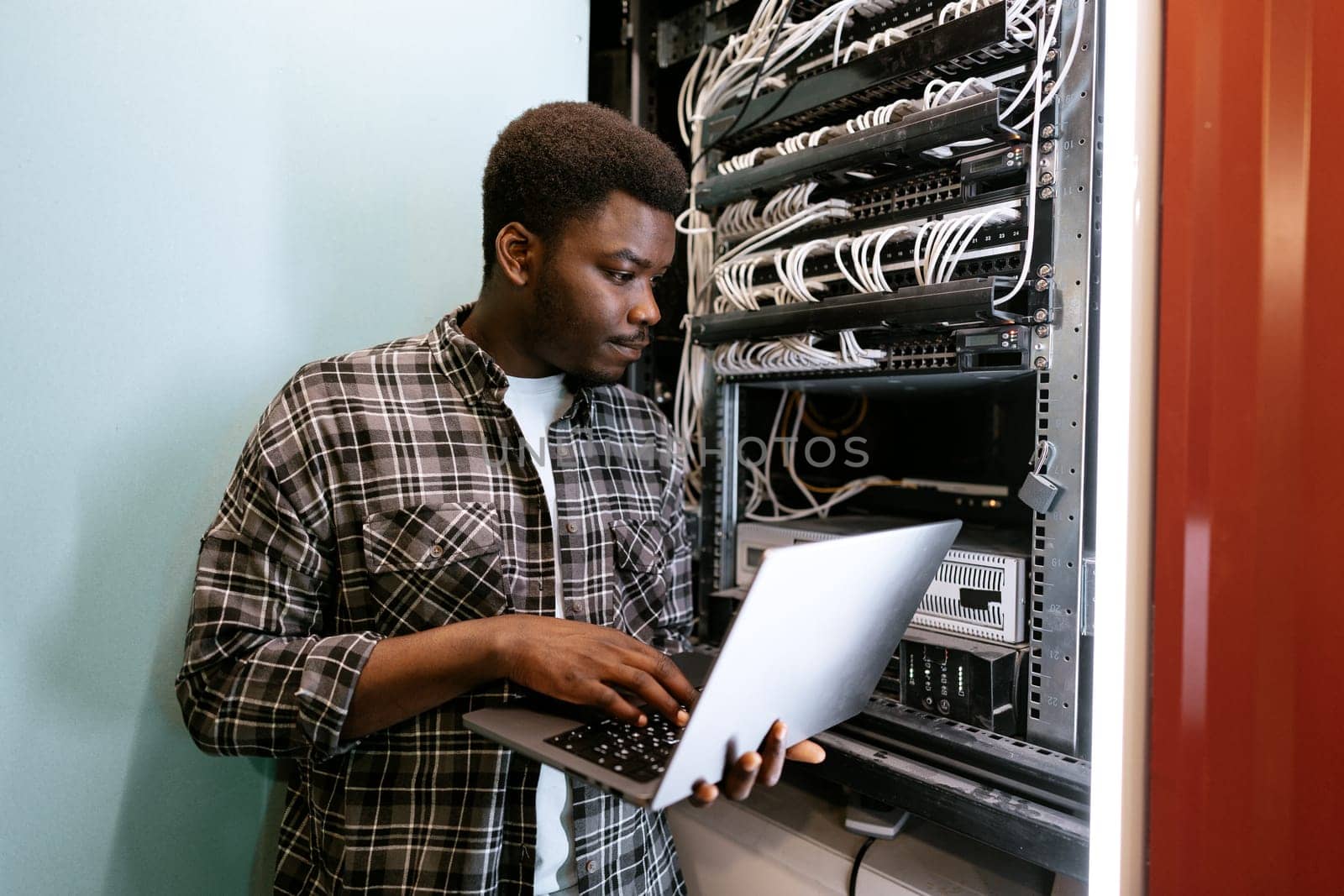A technician is focused on adjusting server settings while holding a laptop in a data center. Cables and servers create a busy yet organized atmosphere in the background.
