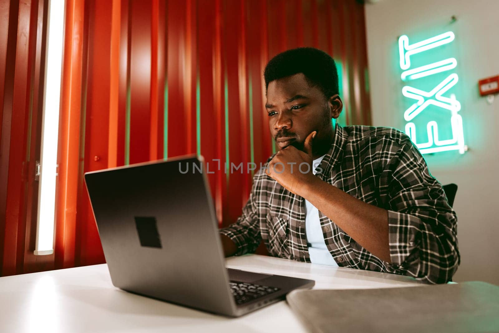 A young man sits at a desk, deeply engaged with his laptop in a stylish workspace. Warm lighting illuminates the environment, enhancing concentration.