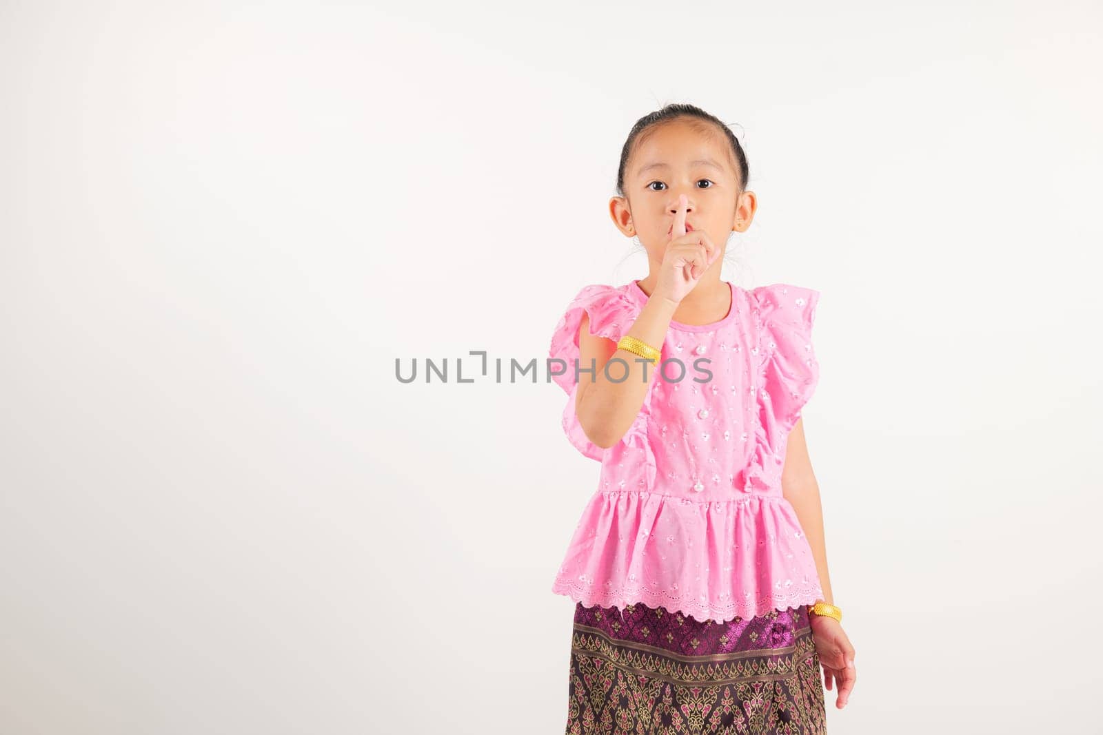 Portrait Thailand kid girl smiling traditional Thai dress costume making finger on lips mouth silent quiet gesture, studio isolated white background, kindergarten child marking silence for stop quiet