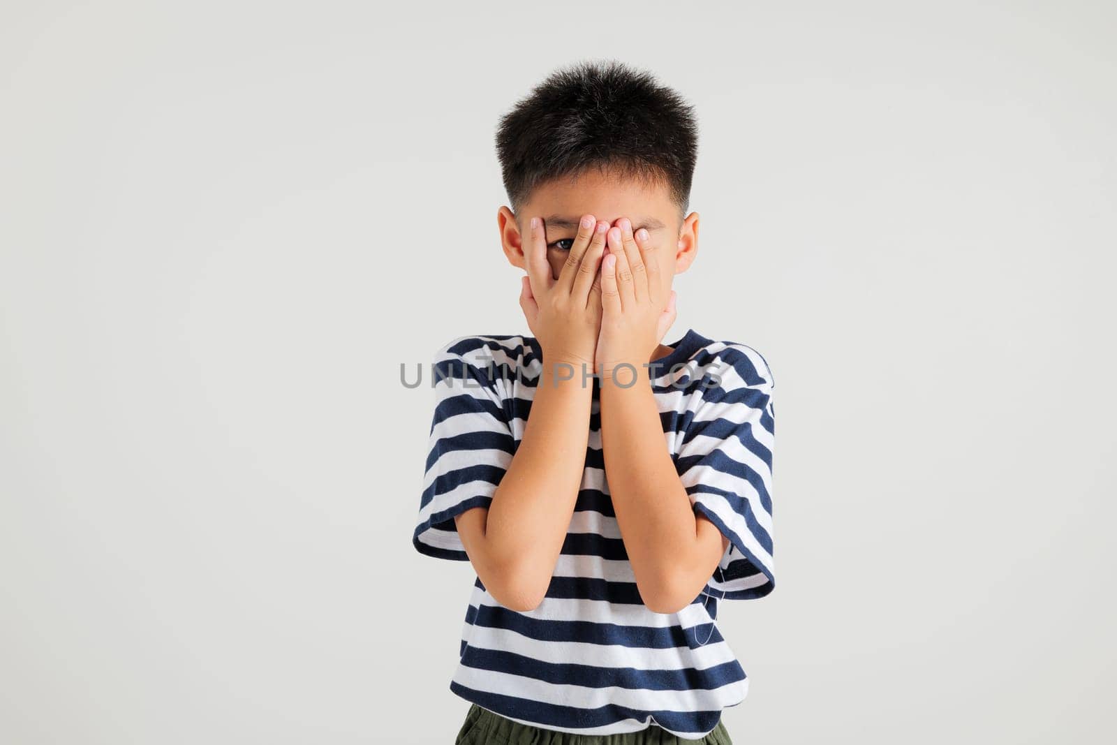 Asian portrait young kid boy in depressed bad mood covering face with hands and peering out with one eye between fingers studio shot isolated on white background, primary child surprised and shocked