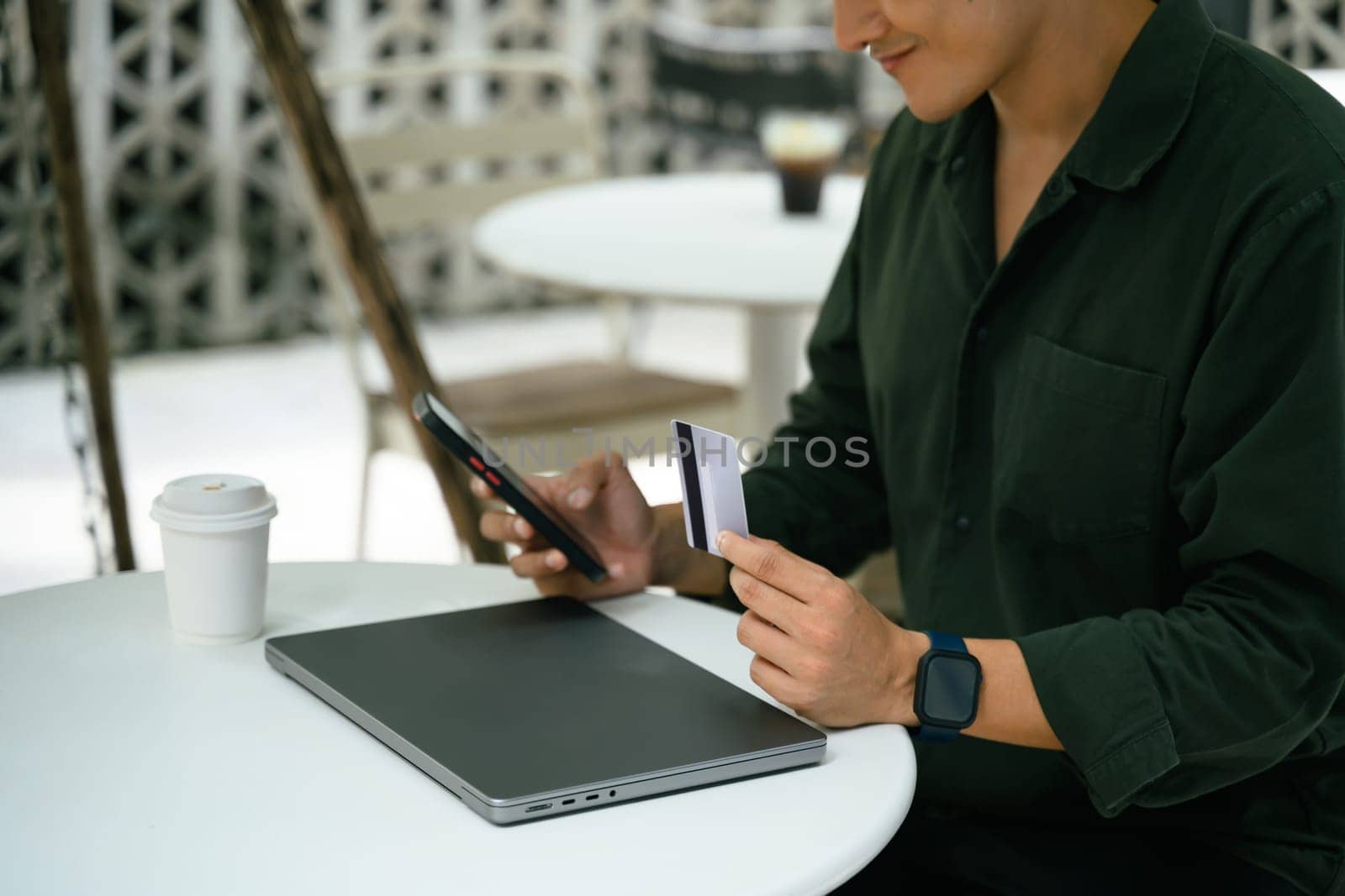 Man holding credit card and using smartphone for an online transaction at an outdoor cafe.
