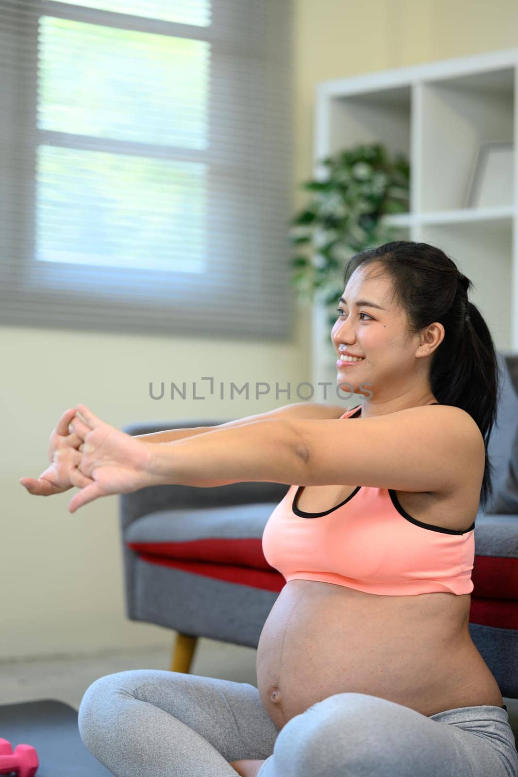Smiling pregnant woman stretches her arms during a gentle workout session at home.