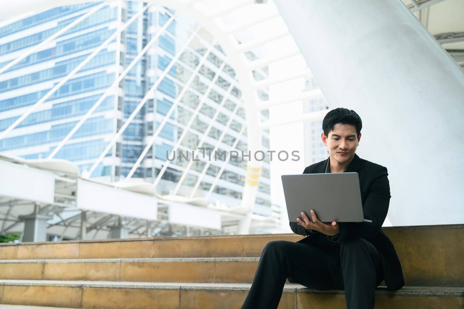 Businessman in formal attire uses laptop while sitting on city steps surrounded by skyscrapers.