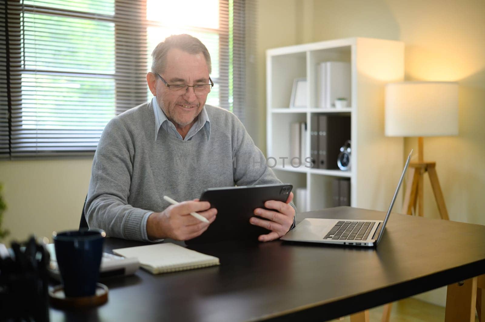 Senior man in sweater and glasses working from home using digital tablet.