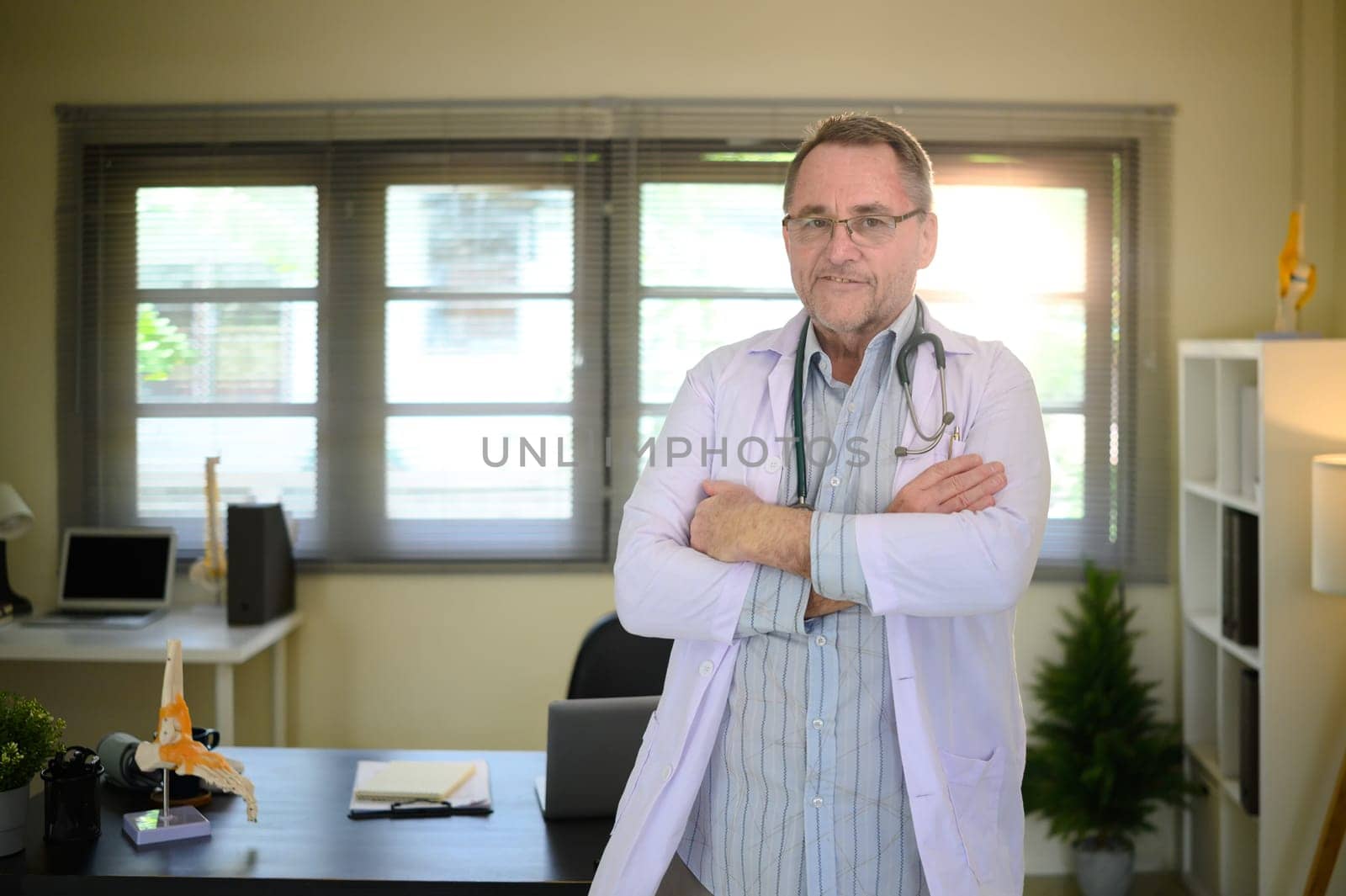 Portrait of experienced male physician with arms crossed in a bright workspace.