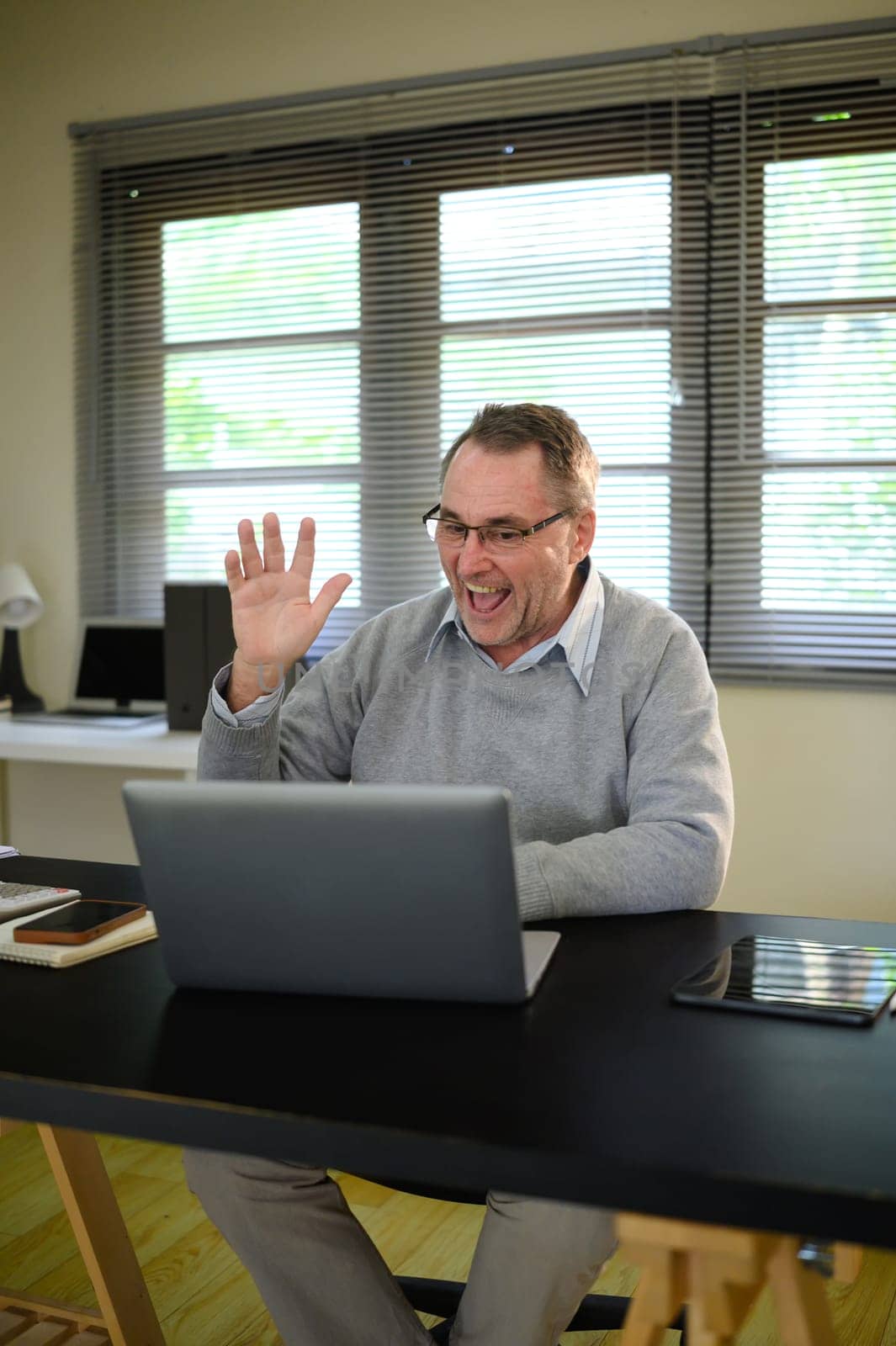 Confident older man enjoys an online conversation from his tidy home office.