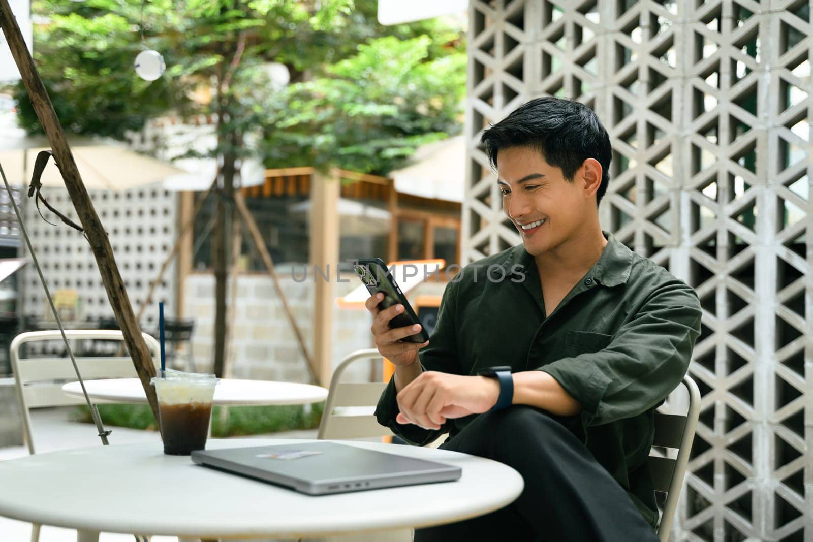Smiling man relaxes at an outdoor cafe and using smartphone beside a closed laptop.