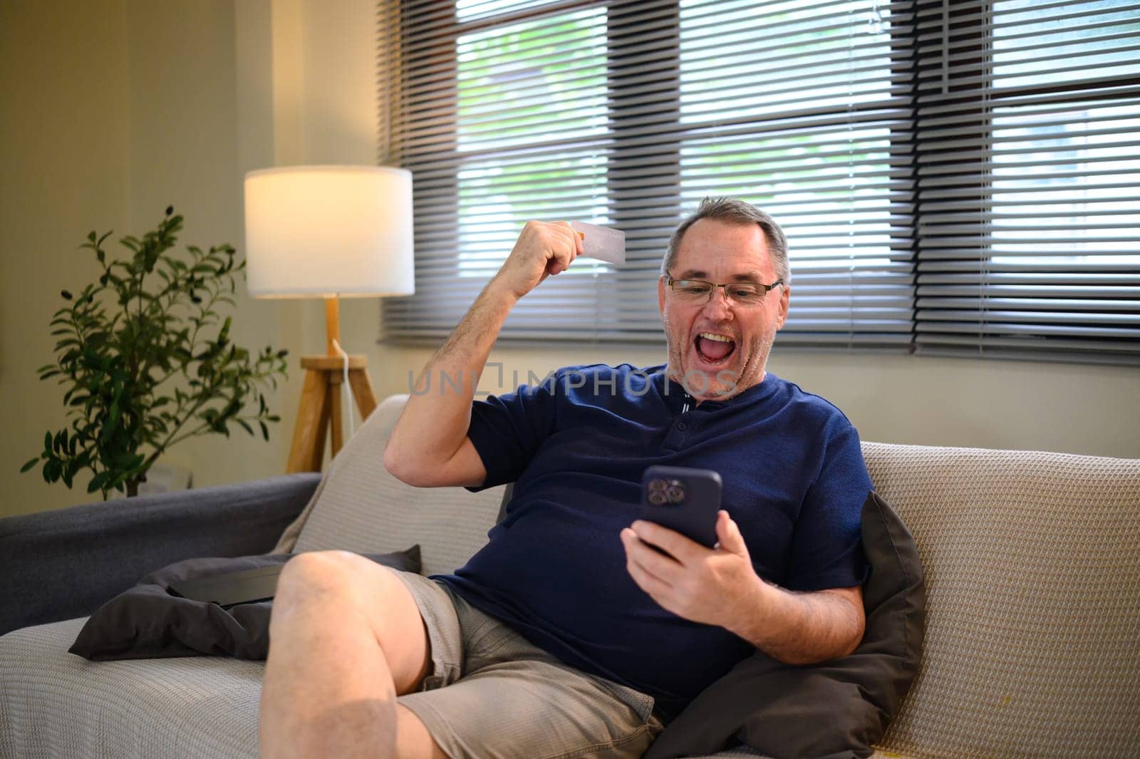 Excited senior man holding credit card and phone, joyful expression after successful transaction.