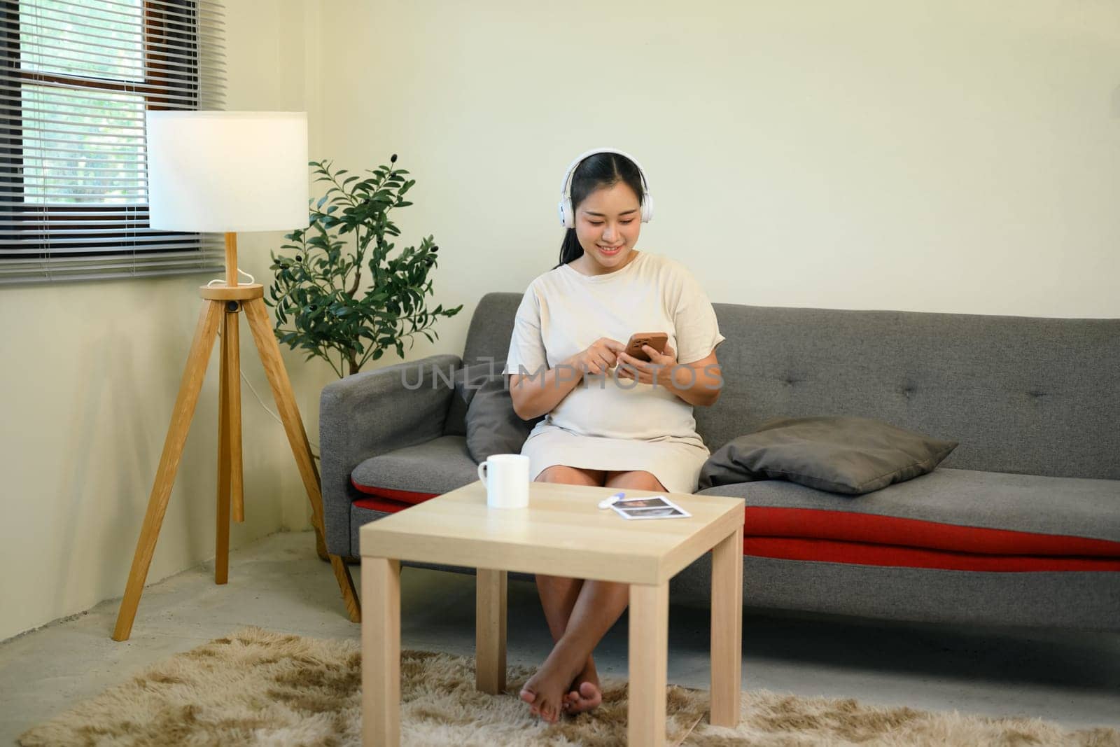 Relaxed pregnant woman sitting on sofa with headphones and using smartphone.