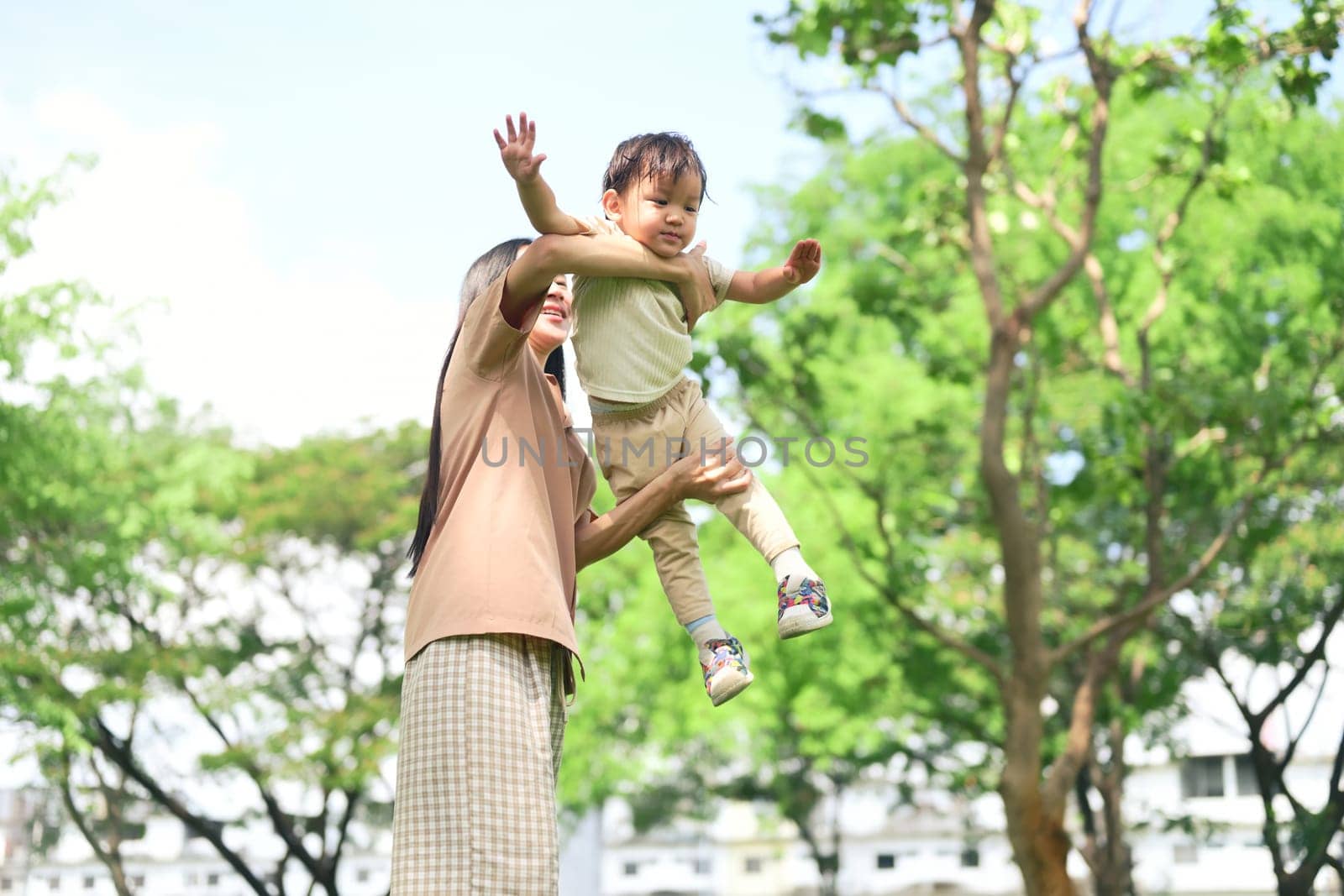Mother smiles as she lifts her young child into the air during a sunny day at the park.