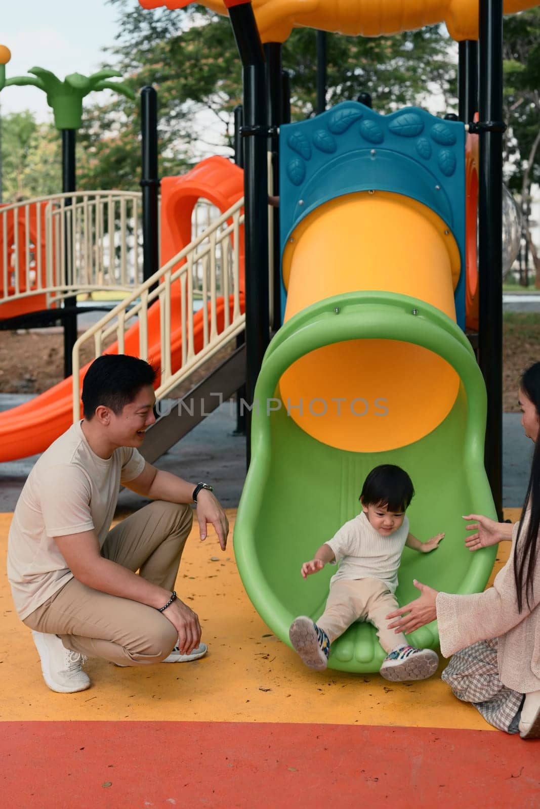 Happy parents watch and cheer as their toddler slides down a colorful slide.