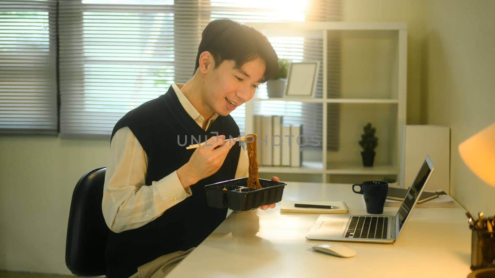 Smiling man eating instant noodles while working at his desk in a cozy, sunlit home office.