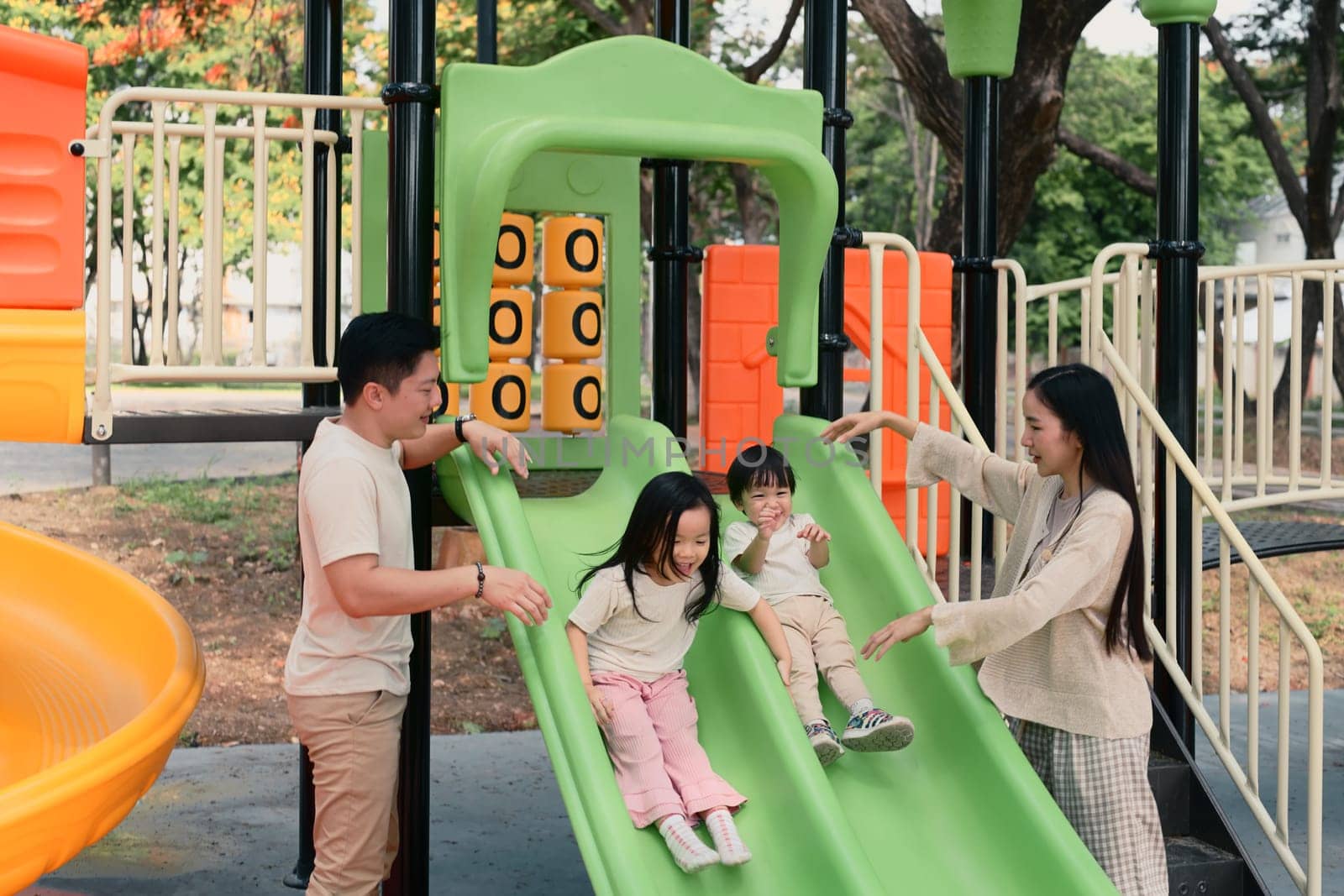 Parents cheer joyfully as two young children enjoy sliding on a playground slide in a sunny park.