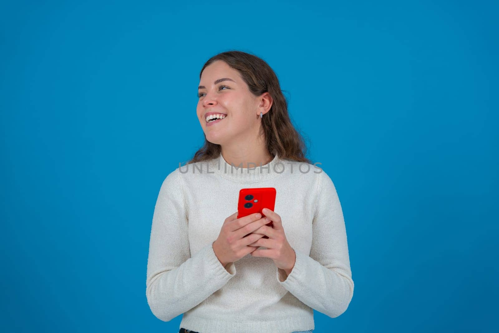 Happy young woman holding a red smartphone while looking to the side with a surprised expression. She stands in front of a bright blue background, expressing excitement and curiosity.