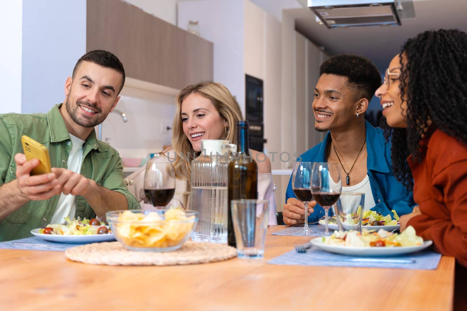 Group of happy multiracial friends sharing snacks and drinks in a cozy modern kitchen. Bright indoor setting, joyful expressions, casual clothing, relaxed and friendly social atmosphere.