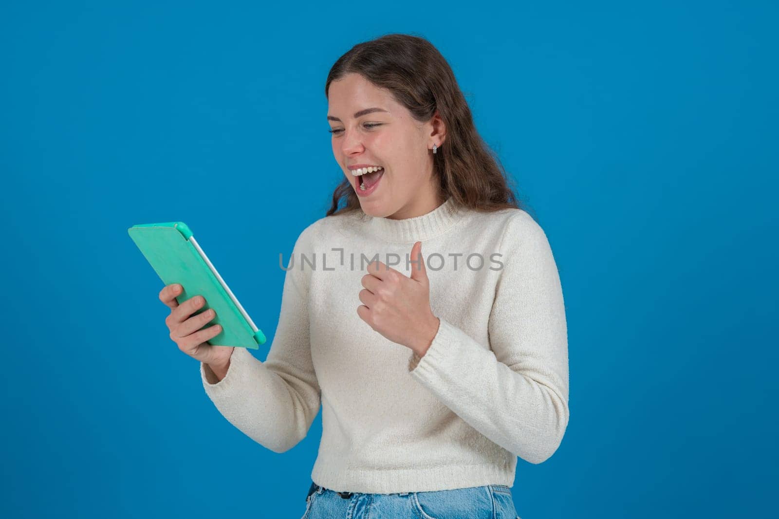 Happy young Caucasian woman holding tablet and showing thumbs up with a cheerful expression. Studio shot on blue background with copy space, ideal for communication or tech concepts.