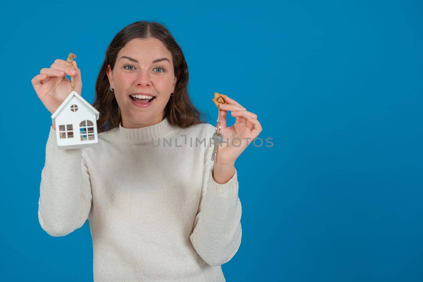 Happy young woman holding a house model and keys, expressing excitement. She wears a white sweater and stands against a blue background. Concept of home ownership, real estate, or investment. Copy space