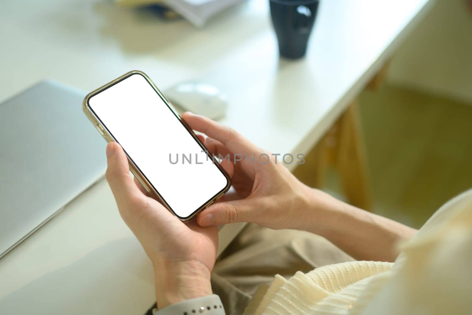 Hands holding smartphone with empty screen sitting in front of laptop at desk.