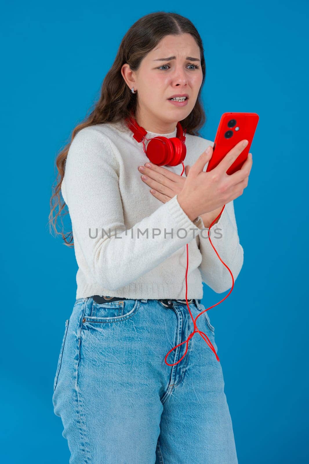Caucasian woman looks shocked while reading bad news on her smartphone. She wears headphones and a white sweater, reacting with concern against a solid blue background.