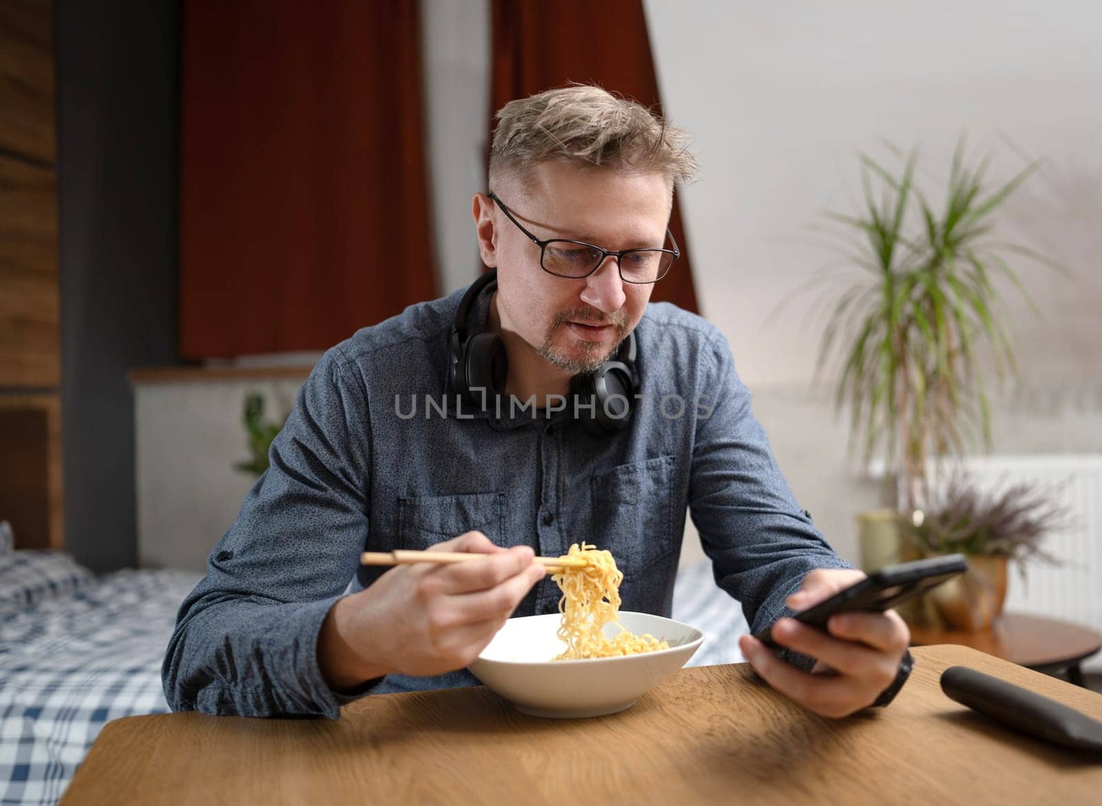 Handsome Man Eating Noodles With Chopsticks And Scrolling Through The News Feed On Smartphone