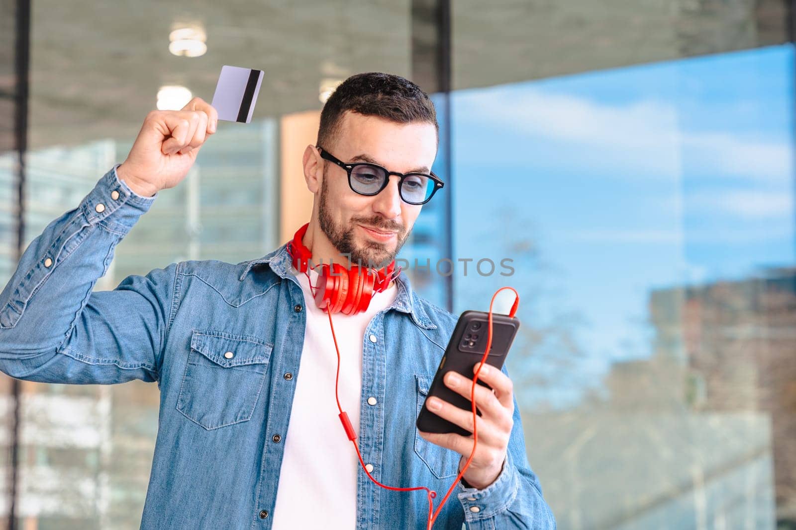 Happy man celebrating a successful online purchase while holding a credit card and smartphone. He smiles with excitement, wearing headphones and sunglasses in a modern urban environment.