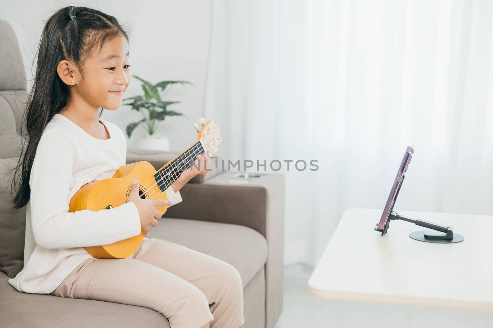A kid girl strums a ukulele while engaging in an online music lesson via a smartphone on a stand. The image portrays tech-savvy, interactive, and joyful home learning experiences.