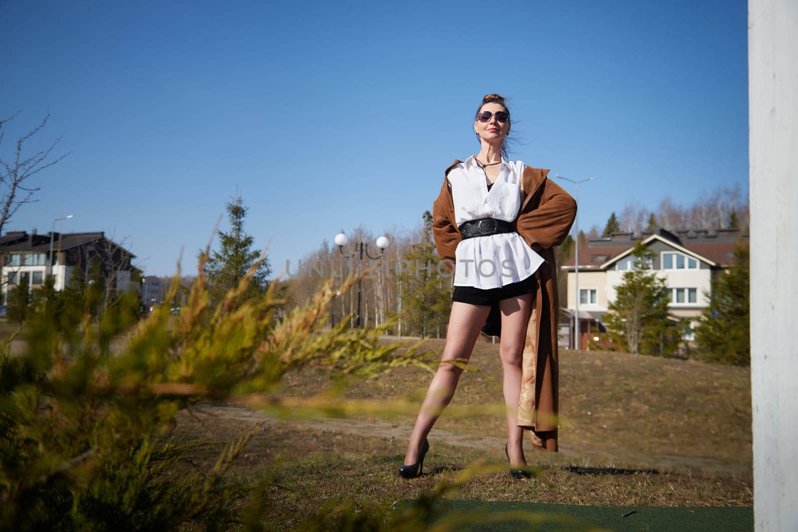 A model showcases a fashionable look with a flowing coat and high heels, standing confidently in a sunny outdoor setting.