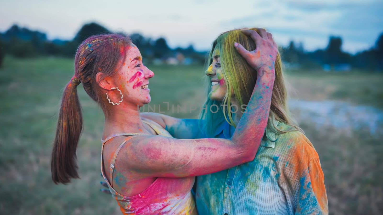 Two school friends, both females, embrace while covered in vibrant powder paint, celebrating the holi festival in a field at sunset