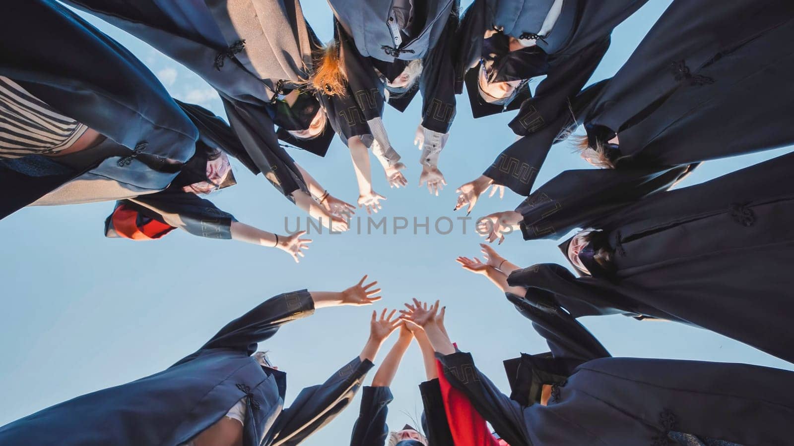 Group of students wearing graduation gowns joining hands forming a circle against blue sky, celebrating their graduation day