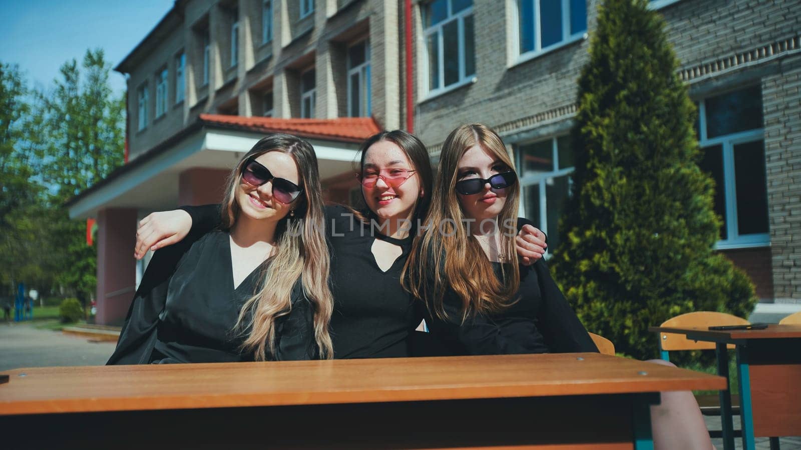 Three cheerful female students wearing sunglasses are posing, embracing each other, sitting at a desk outside their school on a sunny day
