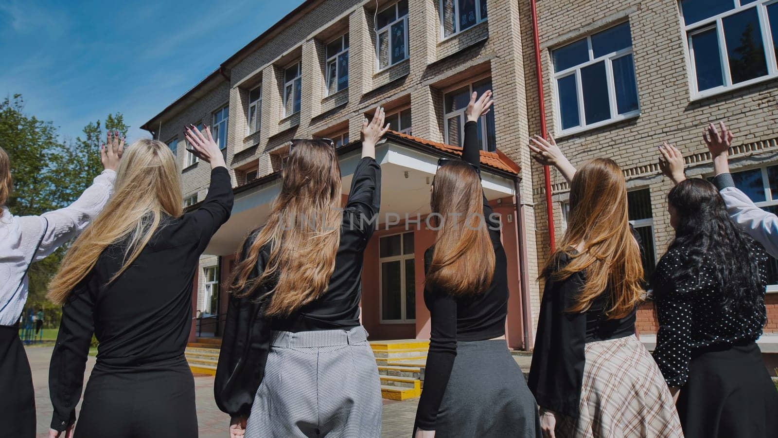 Group of female students joyfully waving goodbye in front of their school building, celebrating their achievements on a sunny day