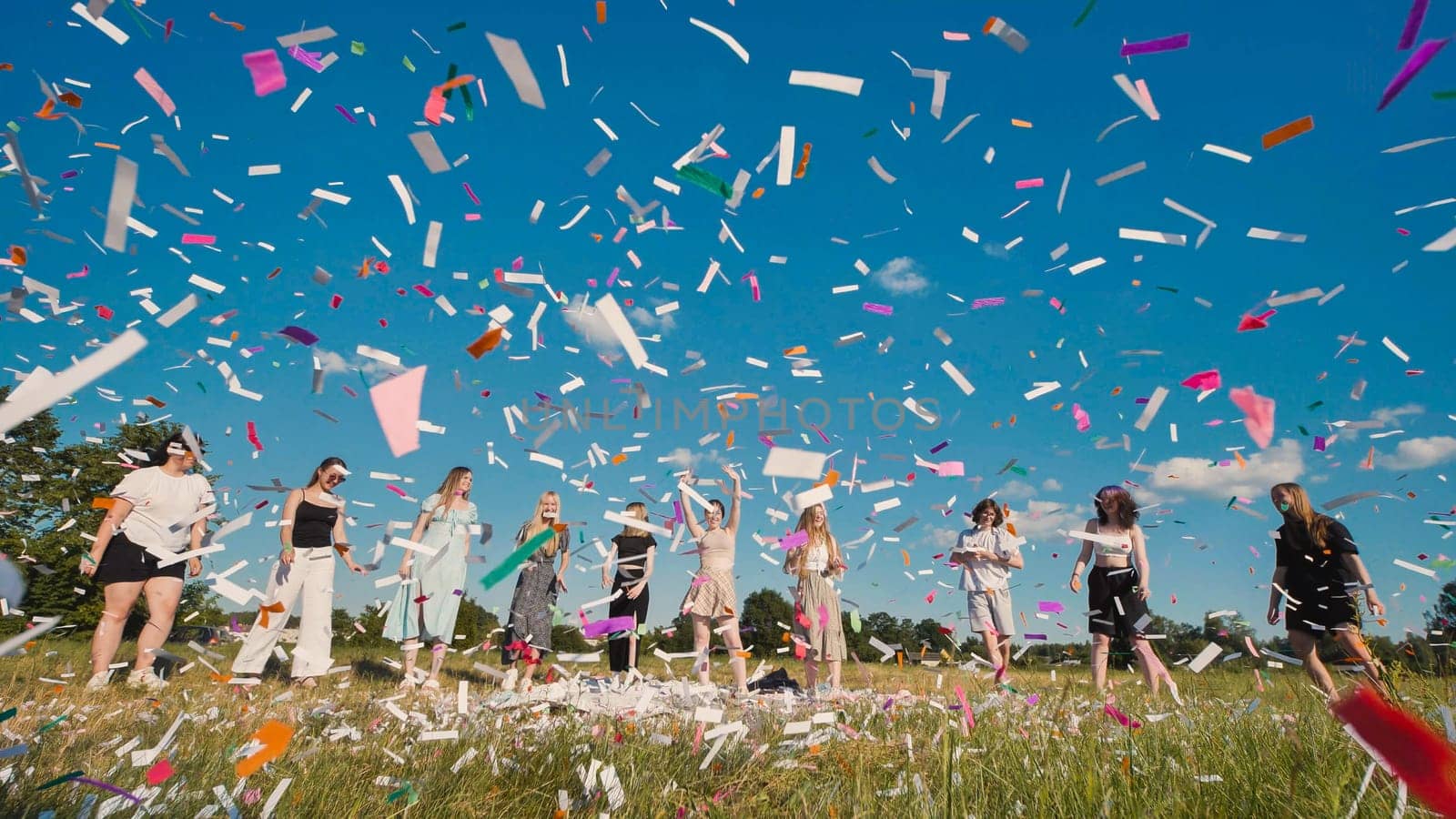 Group of friends joyfully celebrating graduation while throwing colorful confetti in a vibrant field under a bright blue sky