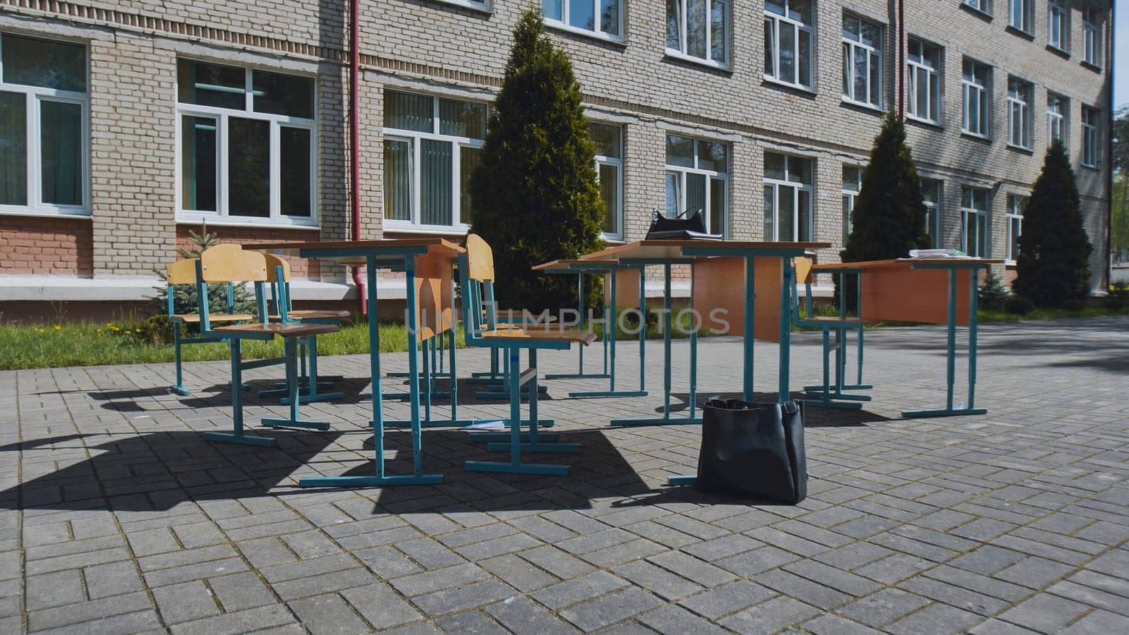 Empty school desks and chairs standing outdoors next to school building during school day with books and bag on the ground