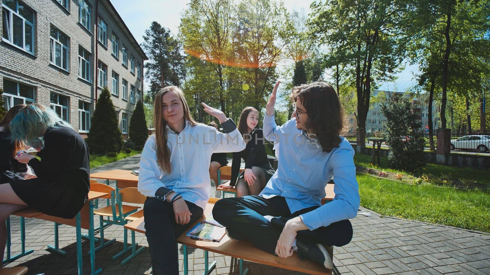 Group of happy students giving high five while sitting on school desks outdoors during break time