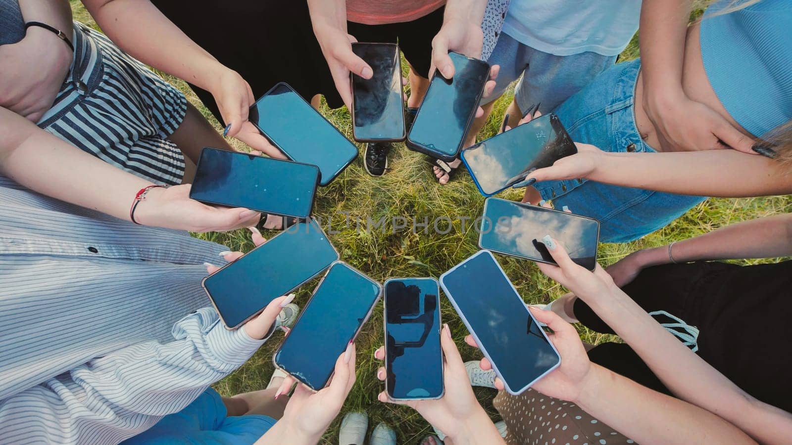 Students holding smartphones creating circle with screens, sharing content and enjoying time together on green grass after school