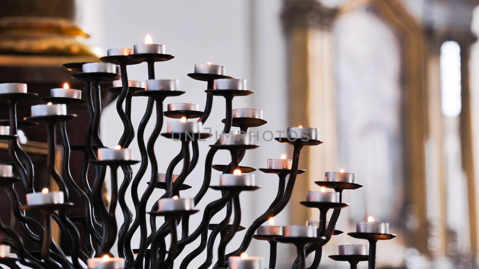Many small burning candles on an elegant wrought iron candelabrum illuminate a church interior