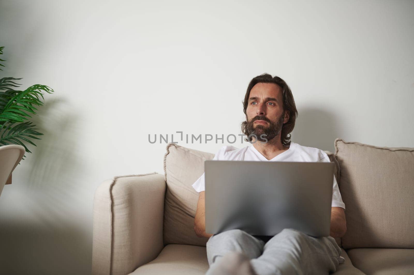 A man is sitting on a comfortable beige couch, working on his laptop, in a stylish and serene minimalist interior setting.