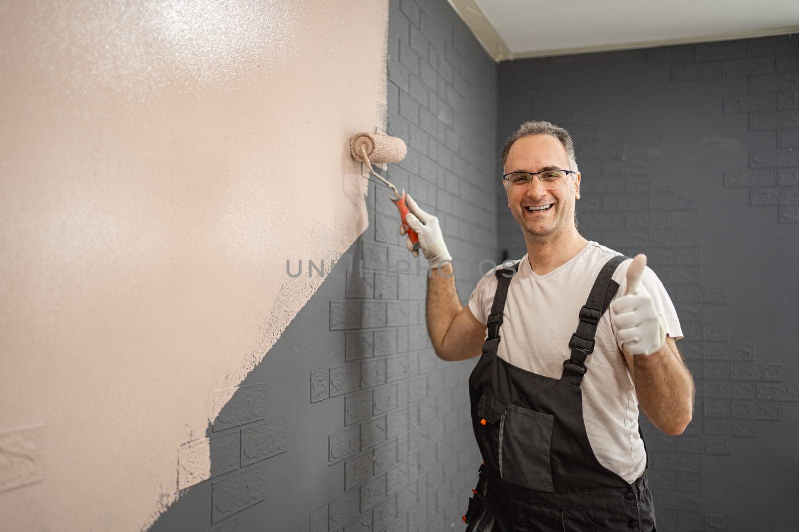 A man is seen standing before a wall, methodically applying paint with a roller. He wears work clothes and holds the roller in his hand, focused on the task at hand.